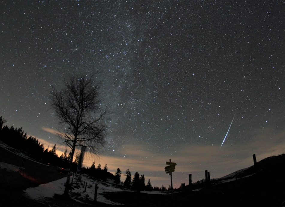 Geminiden-Feuerkugel – aufgenommen von Erwin Filimon am 14. Dez. 2015 um 23:06 Uhr auf der Postalm. Tief am Horizont, beim Masten des Wegweisers ein schwaches Polarlicht
